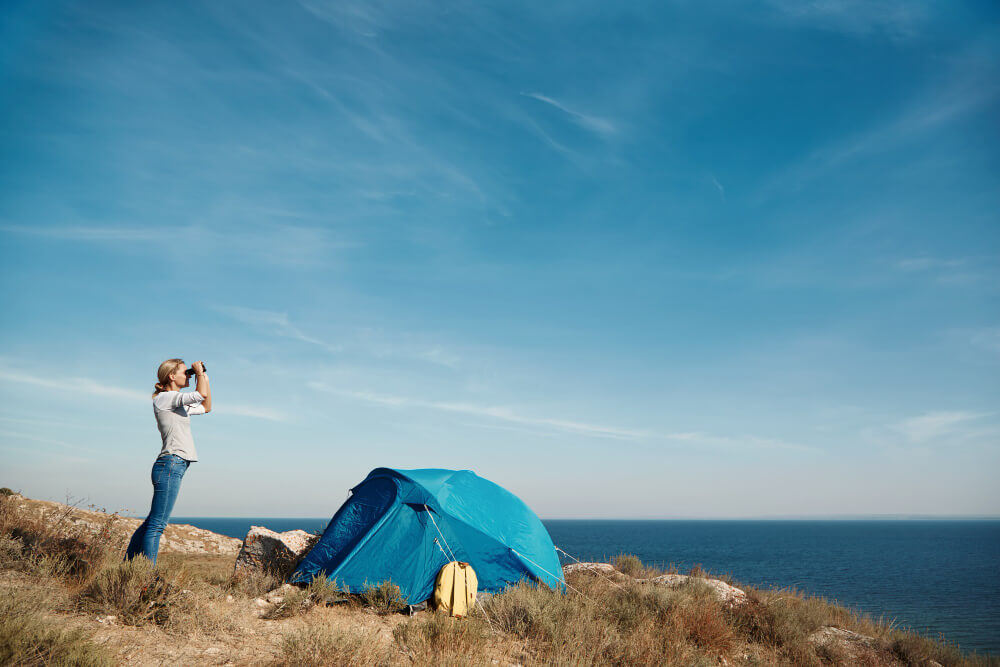 אישה בחוף הים עומדת ליד אוהל ומסתכלת במשקפת